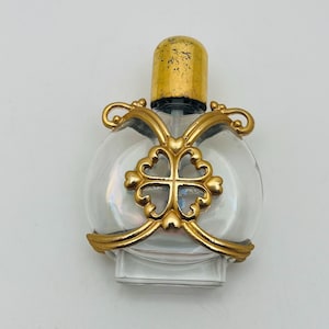 Könnte beinhalten: Eine transparente Parfümflasche aus Glas mit einem goldfarbenen Verschluss und aufwendigem goldfarbenem Filigran. Die Flasche hat einen runden Körper und einen rechteckigen Boden. Das goldene Filigran zeigt ein Kreuzdesign.