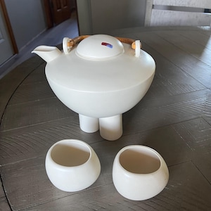 May include: A cream-colored ceramic teapot with a rounded body and three short legs. The lid has a small red and blue accent. Two matching cream-colored teacups are also present. The teapot has a wooden handle.