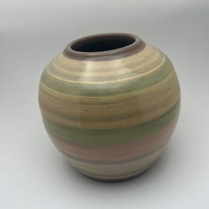 May include: A round ceramic vase with a wide opening and a striped pattern. The vase features bands of beige, green, and peach colors. The rim is a darker brown. The vase is a decorative item.