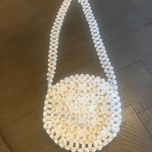 Op de afbeelding: Een ronde handtas gemaakt van witte, bloemvormige kralen die in een dicht, herhalend patroon zijn gerangschikt. De tas heeft een lange, bijpassende schouderband die ook is gemaakt van dezelfde witte, bloemvormige kralen voor een uniform uiterlijk.