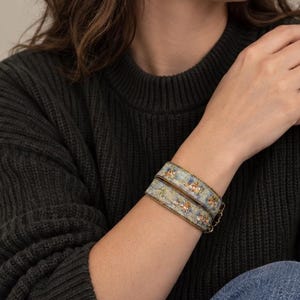 Könnte beinhalten: Ein zweifach gewickeltes Armband mit einem hellblauen Stoffband, das ein zartes Blumenmuster in Gelb- und Grüntönen aufweist, abgerundet durch metallisch goldfarbene Kanten und einen kleinen goldfarbenen Kettenverschluss am Handgelenk.