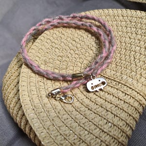 May include: A handmade bracelet with a pink and gray braided design. The bracelet features a silver-colored charm with a cartoon skull design and a silver clasp. The bracelet is resting on a straw hat.