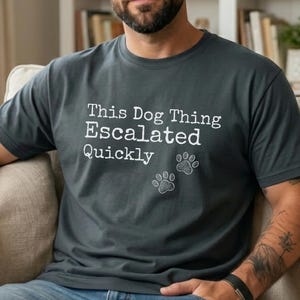Puede incluir: Una camiseta de manga corta en color gris carbón oscuro con el texto blanco "This Dog Thing Escalated Quickly" impreso en el pecho, acompañado de dos pequeñas huellas de perro dibujadas en blanco debajo del texto a la derecha.