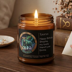 May include: A lit scented soy candle in a glass jar. The candle has a black label with the word "Taurus" and a graphic of a bull. The text "Happy Birthday Shophia, my lovely friend! Love Emma" is also on the label. The candle weighs 9oz.