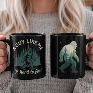May include: Two black ceramic mugs with a Bigfoot design. One mug has the text "A Guy Like Me Is Hard to Find" with a Bigfoot graphic. The other mug features a Bigfoot silhouette with a forest scene.