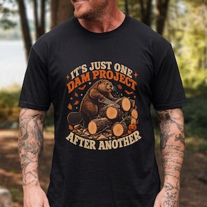 May include: A black short-sleeve t-shirt featuring a graphic of a beaver holding a saw while standing on a pile of logs. The text reads IT'S JUST ONE DAM PROJECT AFTER ANOTHER in orange letters, surrounded by small autumn leaves, bats, and a pumpkin.