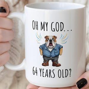 May include: White ceramic mug with the text "OH MY GOD... 64 YEARS OLD?" and a cartoon bulldog wearing a blue shirt and brown boots. The bulldog has a surprised expression and is flanked by blue and yellow lines.