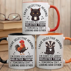 May include: Three ceramic mugs with humorous text and animal illustrations. The mugs feature a black cat, a rooster, and a badger, each holding a cup. The text reads "I have selective hearing" and "I'm sorry you weren't selected today. Tomorrow isn't looking good either."