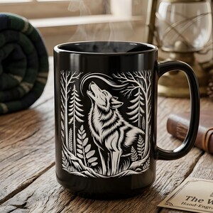 May include: A black ceramic mug featuring a white illustration of a howling wolf in a forest setting. The mug has a large handle and is filled with a steaming beverage. The mug is on a wooden surface, with a rolled-up blanket and pine cones nearby.