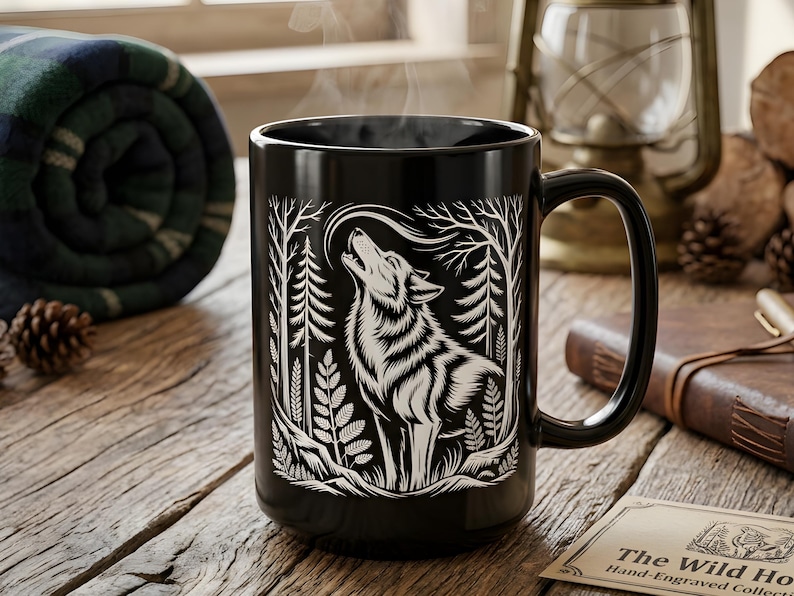 May include: A black ceramic mug featuring a white illustration of a howling wolf in a forest setting. The mug has a large handle and is filled with a steaming beverage. The mug is on a wooden surface, with a rolled-up blanket and pine cones nearby.