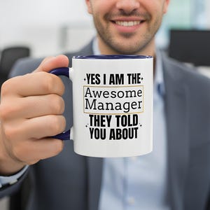 May include: White ceramic mug with a navy blue handle and rim. The mug features the text "YES I AM THE Awesome Manager THEY TOLD YOU ABOUT" in black and gold lettering. The mug is held by a person wearing a gray suit.