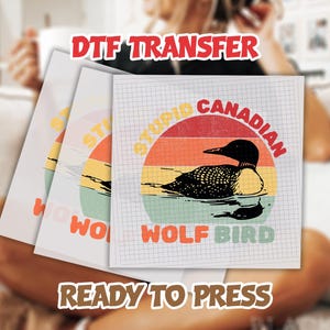 May include: DTF transfer sheets with a graphic of a loon bird in a retro sunset design. The text on the design reads "Stupid Canadian Wolf Bird". The words "DTF TRANSFER" and "READY TO PRESS" are also visible.