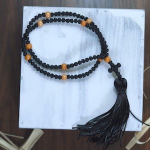 May include: Black rosary with orange beads and a black tassel, resting on a white surface. The rosary features a cross pendant. The background includes wooden elements and decorative straw.