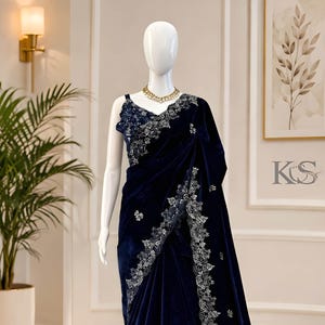 Puede incluir: Un sari de terciopelo azul marino con intrincados bordados plateados. El sari está drapeado sobre un maniquí, mostrando su diseño fluido y sus adornos detallados. Un collar dorado añade un toque de elegancia.