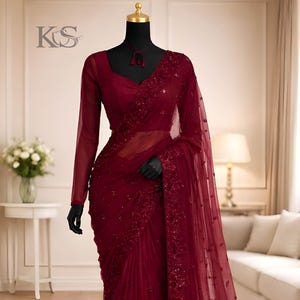 Könnte beinhalten: Ein bordeauxfarbener Sari mit aufwendigen Pailletten- und Perlenverzierungen. Die langärmlige Bluse und der transparente Stoff erzeugen eine elegante Silhouette. Der Sari wird auf einer Schaufensterpuppe präsentiert, um sein Design hervorzuheben. Das Logo "KS" ist sichtbar.