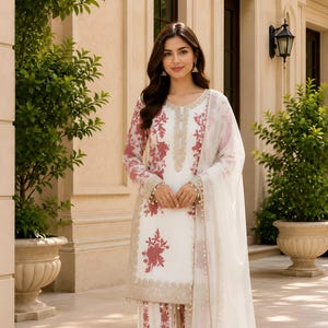 May include: A white three-piece outfit featuring a long-sleeved tunic with a round neckline, matching straight-leg trousers, and a sheer dupatta. The set is decorated with a dark pink floral print and intricate gold embroidery along the edges.
