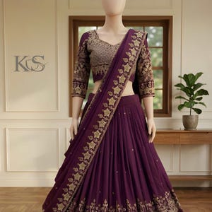 May include: A plum-colored, three-piece Indian formal outfit. The set includes a long skirt, a cropped top with three-quarter sleeves, and a matching scarf. The skirt and scarf feature intricate gold embroidery. The top is embellished with gold sequins.