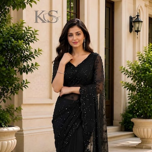 May include: A black saree featuring intricate embroidery and sequin embellishments throughout the fabric. The garment includes a sheer, scalloped border with floral patterns and a matching blouse, showcasing a V-neckline and short sleeves.