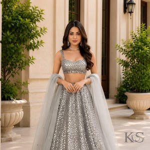 May include: A silver lehenga choli set featuring a sleeveless crop top and a floor-length skirt, both heavily embellished with reflective mirror work. The ensemble includes a sheer, light gray dupatta draped over the arms, creating a cohesive look.
