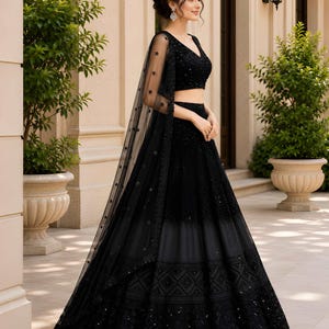 May include: A black lehenga choli set featuring a sleeveless V-neck blouse and a floor-length skirt with intricate embroidery and sequin detailing. A sheer black dupatta with star-shaped embellishments is draped over the shoulder. Text KS is visible.