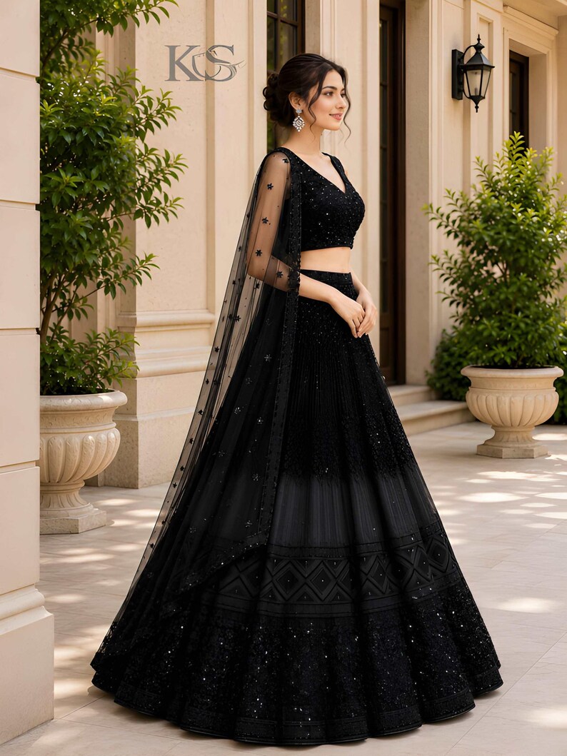 May include: A black lehenga choli set featuring a sleeveless V-neck blouse and a floor-length skirt with intricate embroidery and sequin detailing. A sheer black dupatta with star-shaped embellishments is draped over the shoulder. Text KS is visible.