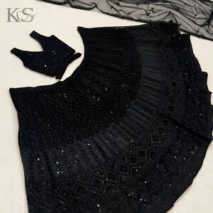 May include: A black three-piece ensemble featuring a sleeveless blouse, a flared skirt, and a sheer dupatta. The skirt and blouse are heavily embellished with intricate black embroidery, geometric patterns, and sequins. The logo KS is visible.