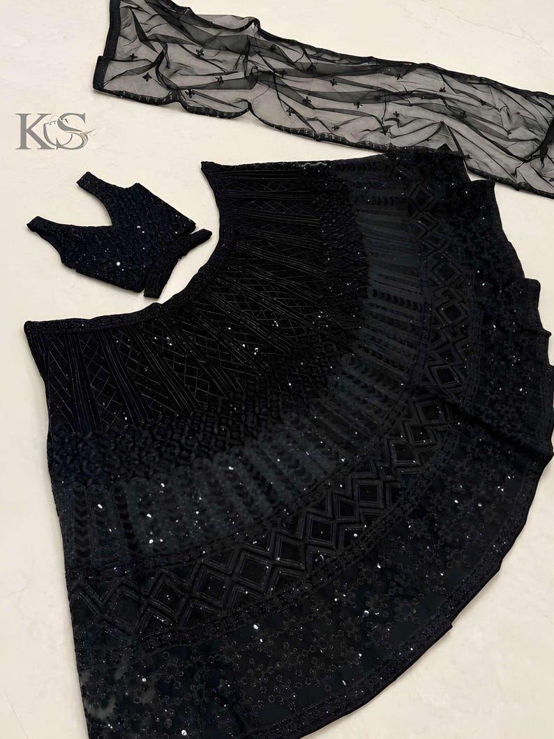May include: A black three-piece ensemble featuring a sleeveless blouse, a flared skirt, and a sheer dupatta. The skirt and blouse are heavily embellished with intricate black embroidery, geometric patterns, and sequins. The logo KS is visible.