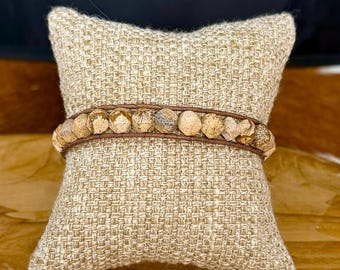 May include: A wrap bracelet featuring natural stone beads in shades of tan and brown. The beads are strung on a brown leather band. The bracelet is displayed on a beige woven pillow.