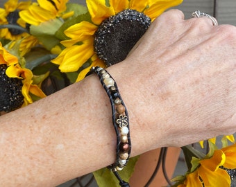 May include: A wrap bracelet featuring dark leather cord, multi-colored beads in shades of brown, cream, and black, and a small metallic flower-shaped charm. The bracelet is constructed with two strands of cord holding the assorted round beads.
