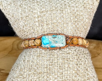 May include: A wrap bracelet featuring a rectangular turquoise stone at the center, surrounded by small, round, tan-colored beads. The bracelet is secured with a brown leather strap. The bracelet is displayed on a beige, textured pillow.