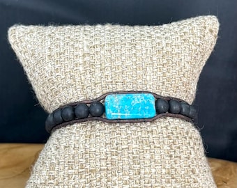 May include: A handcrafted bracelet featuring a rectangular turquoise stone at the center, surrounded by black beads and a dark brown leather band. The bracelet is displayed on a beige woven pillow, showcasing its earthy tones and natural materials.