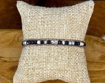 May include: A wrap bracelet with black and white beads and a dark brown leather strap. The bracelet is displayed on a beige woven pillow, showcasing the jewelry's design and texture. The beads are a mix of matte black, white, and silver.