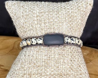 May include: A wrap bracelet featuring a row of speckled, cream-colored beads with black spots. The bracelet is secured by a dark gray rectangular clasp and a brown leather band. The bracelet is displayed on a beige woven pillow.