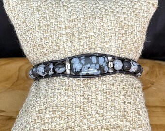 May include: A black and white snowflake obsidian bracelet with a rectangular center stone and smaller round beads. The bracelet is displayed on a beige woven pillow, showcasing the natural stone's unique patterns and colors.
