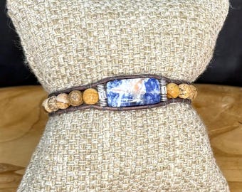 May include: A brown leather wrap bracelet with a rectangular blue and white stone centerpiece. The bracelet features small, round, tan-colored beads and is displayed on a beige woven pillow.