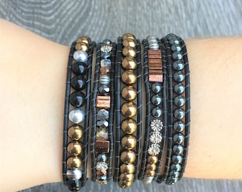 May include: A set of seven black leather wrap bracelets with various beads, including gold, silver, and black. The bracelets feature different patterns and textures, creating a layered and stylish look.