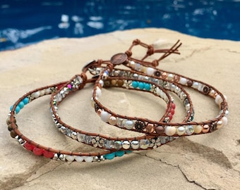 May include: Three leather wrap bracelets with colorful beads, including turquoise, white, and red. The bracelets are layered on a light brown rock.