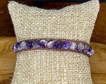 May include: A wrap bracelet featuring amethyst gemstones and a brown leather band. The bracelet is displayed on a small, textured, beige pillow. The amethyst stones are a mix of purple and white.