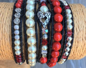 May include: A collection of wrap bracelets featuring various beads and charms. The bracelets include red, white, and silver beads, along with a silver heart charm. The bracelets are wrapped around a tan cylindrical form.