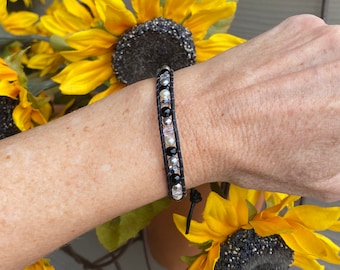 May include: A wrap bracelet featuring two parallel dark leather cords with a central row of alternating black, white, and metallic silver beads, finished with a small black leather tie closure at the end of the beaded section.