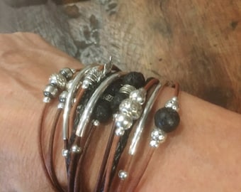 May include: A multi-layered bracelet with brown leather cords, silver beads, and black lava rock beads. The bracelet features a silver charm with a feather design.