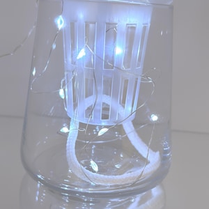 Könnte beinhalten: Ein klares Glas mit einem weißen, geschlitzten Kunststoffkorb im Inneren. Eine Schnur aus kleinen weißen LED-Leuchten ist um den Korb und in das Glas gewickelt. Ein weißes Seil erstreckt sich vom Korb bis zum Boden des Glases.