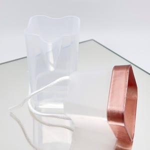 Könnte beinhalten: Ein durchscheinender weißer Kunststoffbehälter mit einer sternförmigen Öffnung und einem metallischen roségoldenen Rand. Eine dünne weiße Kordel ist an der Seite des Behälters befestigt, der eine glatte, halbtransparente Oberfläche und eine geometrische Form aufweist.
