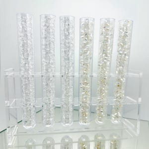 Könnte beinhalten: Ein klarer Acryl-Präsentationsständer mit sechs transparenten Glasröhren, die mit funkelnden Kristallen gefüllt sind. Die Kristalle variieren in der Farbe, von klar bis irisierend. Die Röhren sind in zwei Reihen angeordnet, mit kleineren Röhren darunter.