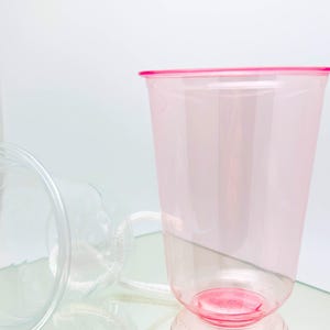 Może przedstawiać: Przezroczysty plastikowy kubek z różową obręczą i podstawą. Kubek jest częściowo wypełniony jasno różowym płynem. Drugi przezroczysty kubek leży z boku w tle.