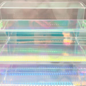 Könnte beinhalten: Drei transparente Acryl-Ausstellungsboxen, die übereinander gestapelt sind. Die Boxen haben schillernde Kanten, die einen Regenbogen von Farben reflektieren. Die Boxen sind rechteckig mit einem Rand an der Oberkante.