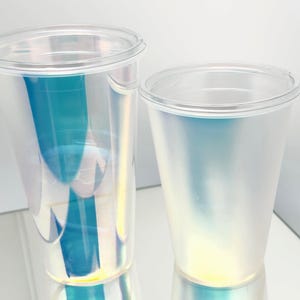 Könnte beinhalten: Zwei transparente Plastikbecher mit Deckel. Die Becher haben eine schillernde Oberfläche, die blaue, grüne und gelbe Farbtöne reflektiert. Die Becher sind leer und scheinen aus leichtem Material zu bestehen. Die Becher eignen sich für kalte Getränke.