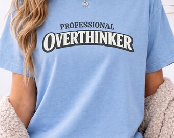 May include: A light blue short-sleeve t-shirt featuring the text "PROFESSIONAL OVERTHINKER" printed in a dark, bold, arched font across the chest. The shirt has a classic crew neckline and a relaxed fit, made of a solid-colored cotton fabric.