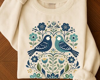 May include: Cream-colored sweatshirt featuring a symmetrical design of two birds facing each other, surrounded by blue and teal floral elements. The birds are in shades of blue with white detailing. The design is centered on the front of the long-sleeved top.