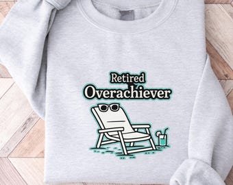 May include: Light gray sweatshirt with the words "Retired Overachiever" in a black and teal font. The graphic features a white lounge chair with sunglasses and a drink with a straw.